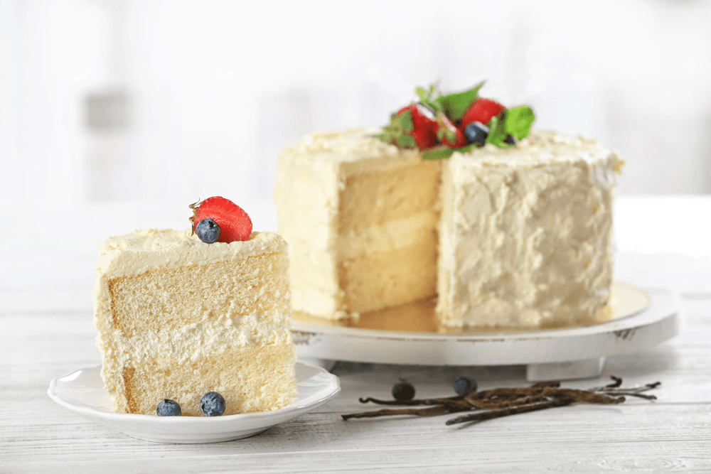 A slice of vanilla cake topped with berries, alongside a whole cake on a display stand.