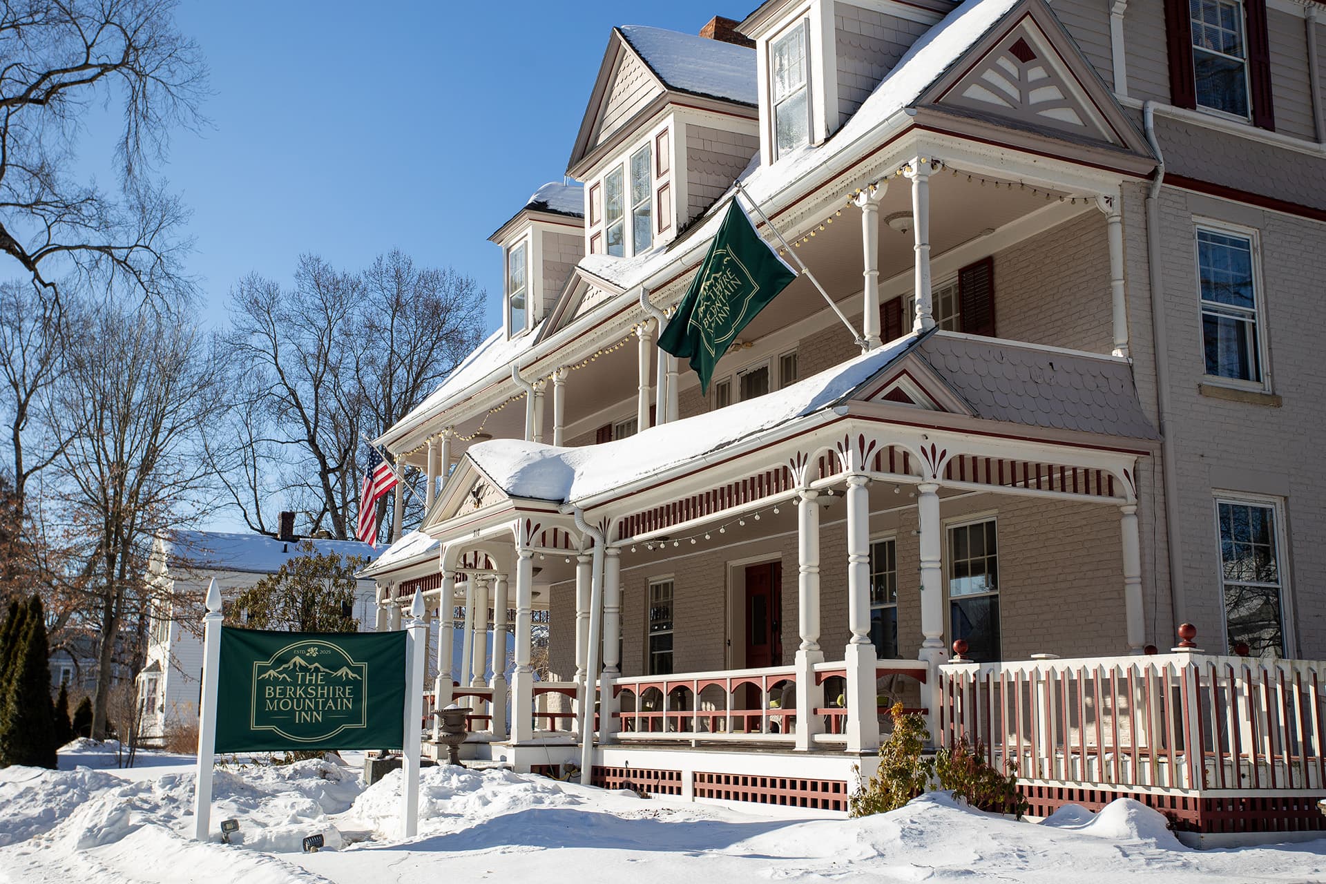 A historic inn with a porch, surrounded by snow and trees, featuring American flags and a welcoming sign.