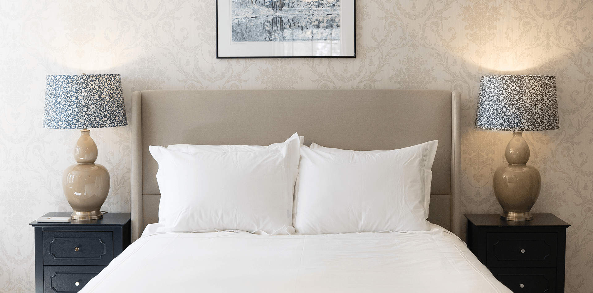 A neatly made bed with white pillows is flanked by two lamps on dark nightstands, under a decorative wall.